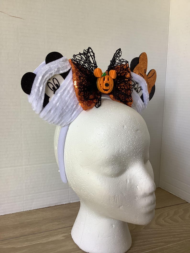 Op de afbeelding: Een wit hoofdband met een zwart en oranje strikje, een wit, pluche Mickey Mouse-oor met een mummie en een oranje Mickey Mouse-oor met een pompoen erop.