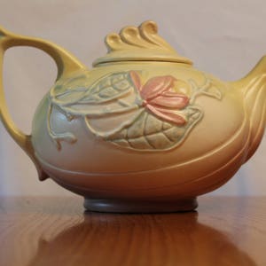 May include: A yellow teapot with a floral design. The teapot has a lid and a spout. The floral design is in shades of pink, green, and yellow.