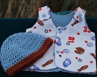 May include: A sleeveless vest with a white background featuring a baseball-themed print of bats, balls, gloves, and red stars, secured with light blue snaps, paired with a light blue and brown crocheted beanie hat with a textured stitch pattern.