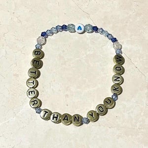 May include: A bracelet with gold-coloured letter beads spelling "BETTER THAN YOU KNOW". The bracelet also features blue and clear beads, with a white bead with a blue heart.