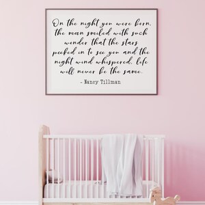 Op de afbeelding: Een wit ingelijst print met een zwart-wit citaat over de nacht dat een baby werd geboren. Het citaat luidt "On the night you were born, the moon smiled with such wonder that the stars peeked in to see you and the night wind whispered, life will never be the same." - Nancy Tillman. De print hangt aan een roze muur boven een wit wiegje.