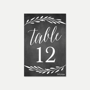 May include: Black chalkboard table number with white calligraphy that says "table 12" with white leafy branches at the top and bottom. The text "holly designs" is at the bottom right corner.