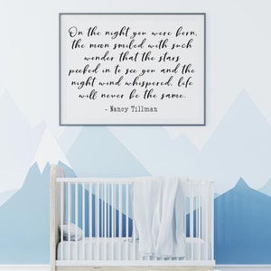 Op de afbeelding: Een wit babybedje met een wit dekentje in een kinderkamer met een blauwe en witte bergmuur. Een ingelijst print met de quote "On the night you were born, the moon smiled with such wonder that the stars peeked in to see you and the night wind whispered, life will never be the same. - Nancy Tillman" hangt aan de muur.