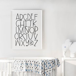 May include: A white framed print with black lettering of the alphabet, A through Z, and an ampersand. The print is hanging on a white wall above a white crib with a blue and white patterned blanket.