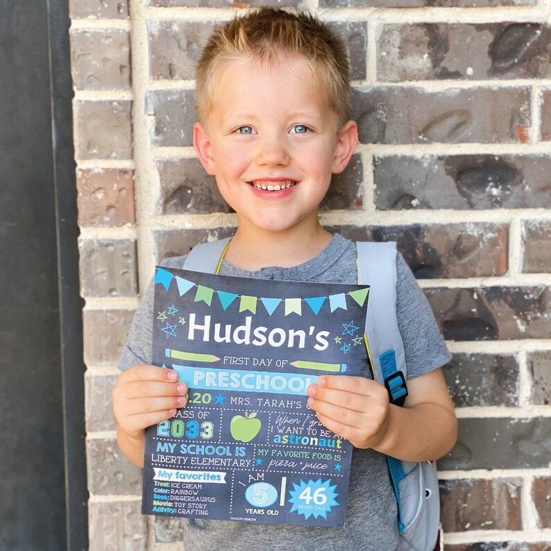 K&ouml;nnte beinhalten: Ein Kind h&auml;lt ein Tafelschild mit der Aufschrift "Hudson's First Day of Preschool". Das Schild enth&auml;lt Details &uuml;ber die Schule des Kindes, seine Lieblingsdinge und eine Gr&ouml;&szlig;e von 117 cm. Das Kind tr&auml;gt ein graues T-Shirt und einen Rucksack.