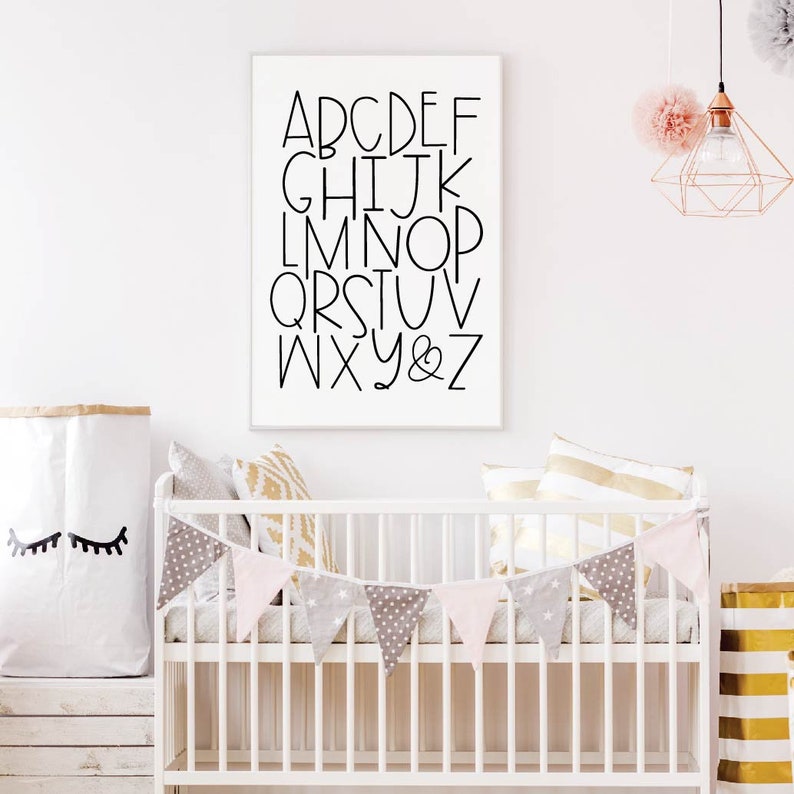 May include: A white framed print with black lettering of the alphabet, A through Z, and an ampersand. The print is hanging on a white wall above a white crib with a pink and gray bunting.