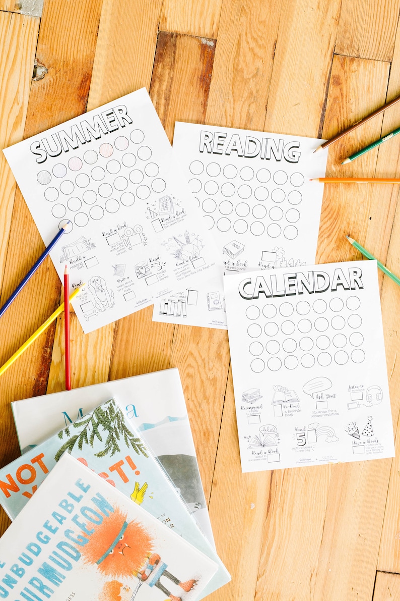 Op de afbeelding: Een set printbare leeskaarten met cirkels om in te kleuren om leesdoelen bij te houden. De kaarten zijn getiteld "Summer", "Reading" en "Calendar". De kaarten liggen op een houten oppervlak met boeken en kleurpotloden.