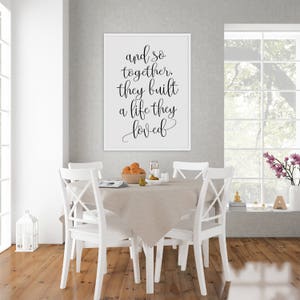 May include: A white framed print with black calligraphy that reads "and so together, they built a life they loved". The print is hanging on a gray wall above a white dining table with four white chairs. There is a bowl of fruit on the table.