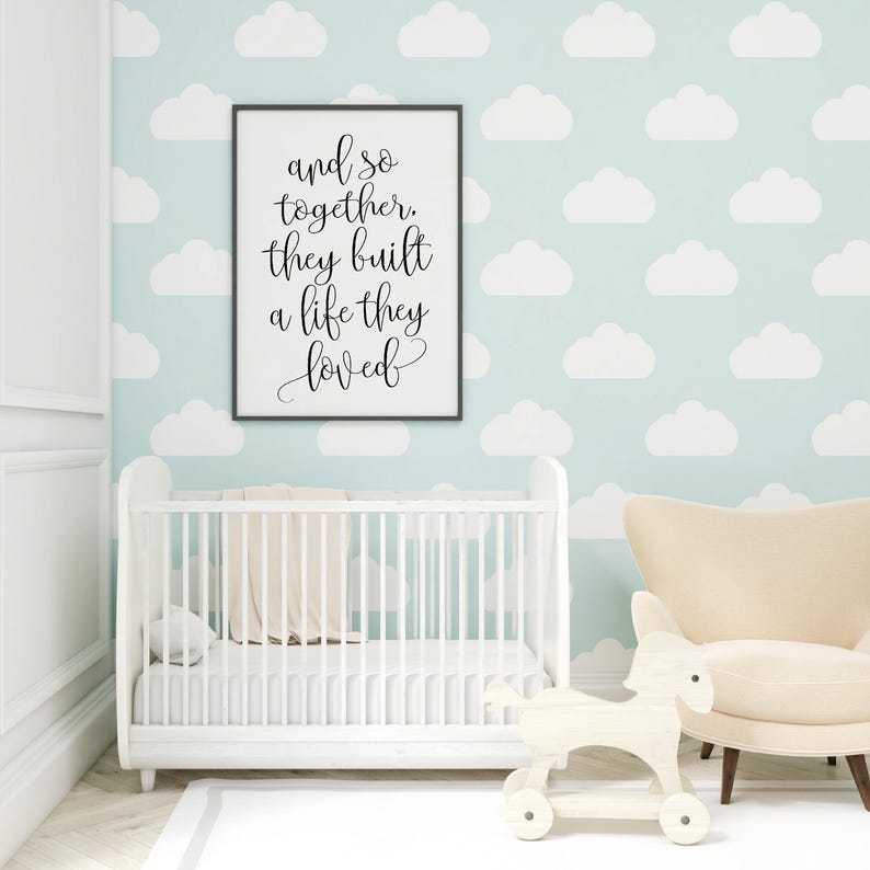 May include: A white crib with a white blanket and a black framed print with the text "and so together they built a life they loved" hanging on a light blue wall with white cloud patterns.