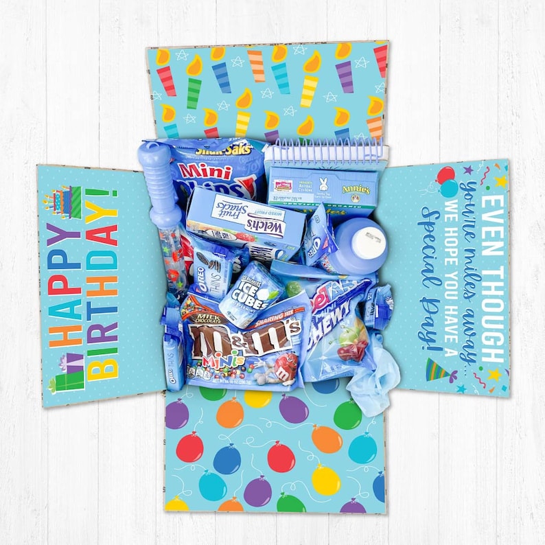 May include: A blue and white gift box filled with various blue and colorful snacks and treats. The box has a happy birthday message on the front and a message that says "Even though you're miles away, we hope you have a special day!" on the side.