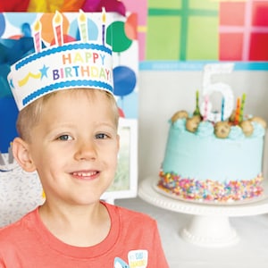 Puede incluir: Un ni&ntilde;o peque&ntilde;o con una corona de papel de 'Feliz Cumplea&ntilde;os' de colores sonr&iacute;e a la c&aacute;mara. Est&aacute; de pie frente a un pastel de cumplea&ntilde;os decorado con glaseado azul y chispas de colores.