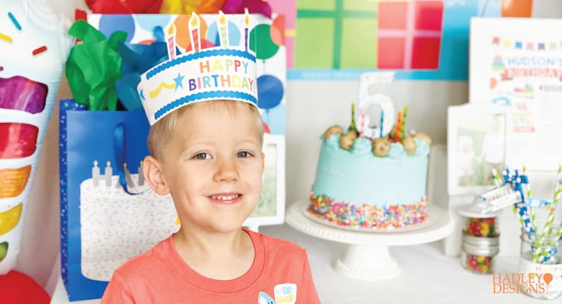 Puede incluir: Un ni&ntilde;o peque&ntilde;o con una corona de papel de 'Feliz Cumplea&ntilde;os' de colores sonr&iacute;e a la c&aacute;mara. Est&aacute; de pie frente a un pastel de cumplea&ntilde;os decorado con glaseado azul y chispas de colores.