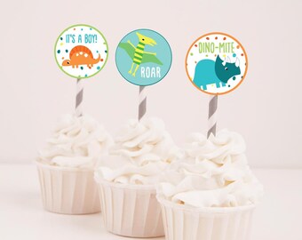 Könnte beinhalten: Drei Cupcakes mit weißem Frosting, verziert mit runden Cupcake-Toppern zum Thema Dinosaurier. Die Topper zeigen einen grünen Dinosaurier mit dem Text "Roar", einen blauen Dinosaurier mit dem Text "Dino-mite" und einen orangefarbenen Dinosaurier mit dem Text "It's a Boy!"
