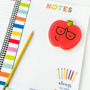 Pode incluir: Um caderno branco com uma capa listrada colorida e uma encaderna&ccedil;&atilde;o em espiral. O caderno tem a palavra "NOTES" escrita em letras coloridas no topo. Um biscoito de ma&ccedil;&atilde; vermelho com um caule verde e &oacute;culos pretos est&aacute; no caderno. O biscoito tem um rosto sorridente. O caderno tamb&eacute;m tem uma se&ccedil;&atilde;o com o texto "always do your best" e uma fileira de l&aacute;pis de cor.