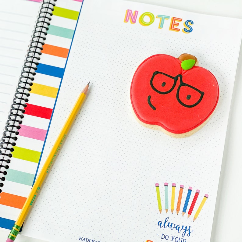 Pode incluir: Um caderno branco com uma capa listrada colorida e uma encaderna&ccedil;&atilde;o em espiral. O caderno tem a palavra "NOTES" escrita em letras coloridas no topo. Um biscoito de ma&ccedil;&atilde; vermelho com um caule verde e &oacute;culos pretos est&aacute; no caderno. O biscoito tem um rosto sorridente. O caderno tamb&eacute;m tem uma se&ccedil;&atilde;o com o texto "always do your best" e uma fileira de l&aacute;pis de cor.
