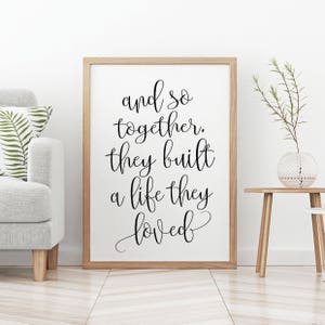 May include: A framed print with a black and white quote that reads "and so together, they built a life they loved". The print is in a wooden frame and is hanging on a white wall.