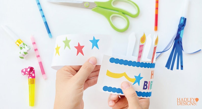 Puede incluir: Un gorro de cumplea&ntilde;os de papel blanco con un dise&ntilde;o de pastel azul y amarillo, tres velas y una estrella. El gorro est&aacute; decorado con estrellas verdes, azules, rojas y amarillas.