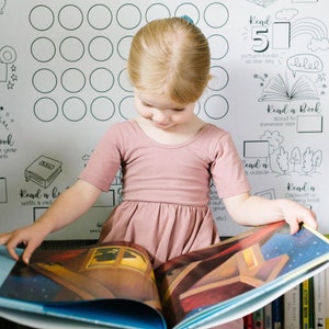Op de afbeelding: Een jong meisje in een roze jurk leest een boek. Het boek is open en de pagina's zijn gevuld met kleurrijke illustraties. Het meisje zit voor een witte poster met een lijst van leesuitdagingen. De poster heeft zwart-wit illustraties van boeken, een kalender en een filmcamera.