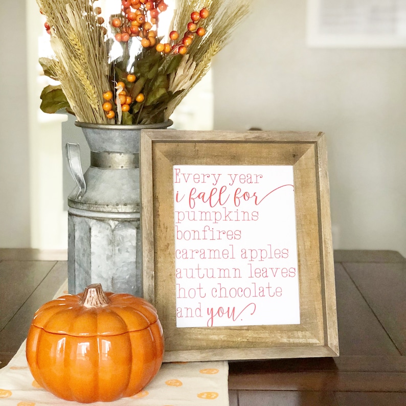 May include: A framed print with the text "Every year i fall for pumpkins bonfires caramel apples autumn leaves hot chocolate and you." The print is on a wooden table with a ceramic pumpkin and a vase of dried flowers.