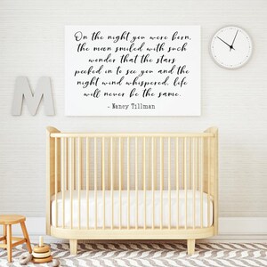 Op de afbeelding: Een wit ingelijst print met een zwart-wit citaat over de nacht dat een baby werd geboren. Het citaat luidt "On the night you were born, the moon smiled with such wonder that the stars peeked in to see you and the night wind whispered, life will never be the same. - Nancy Tillman". De print hangt aan een witte muur boven een houten wieg.
