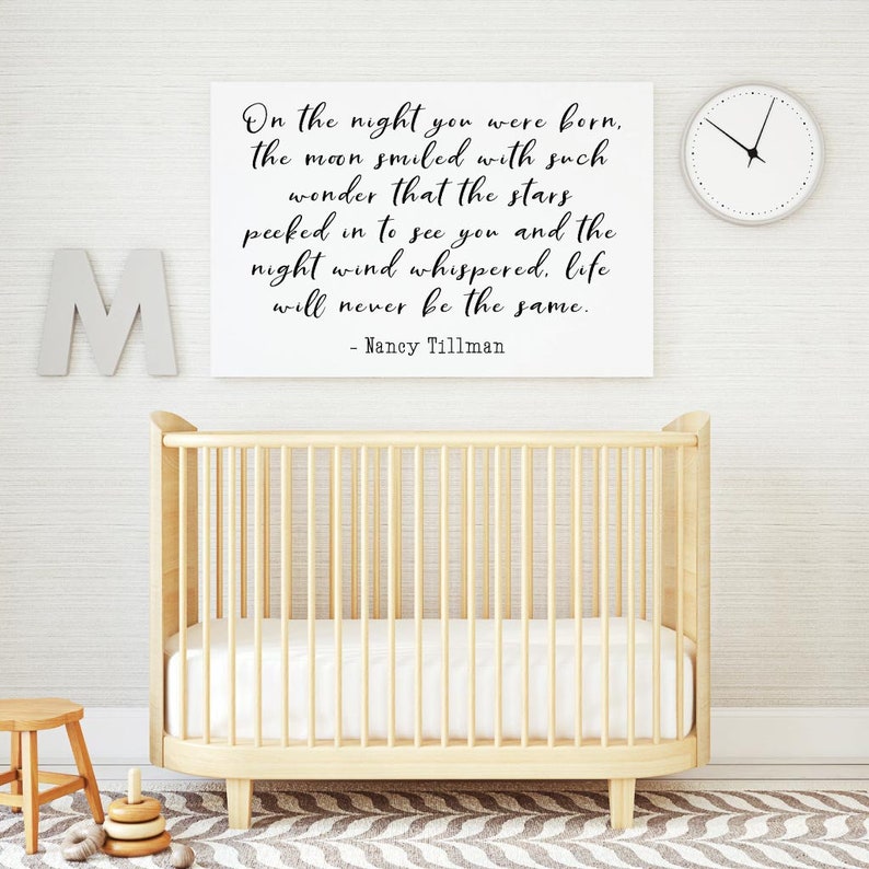 Op de afbeelding: Een wit ingelijst print met een zwart-wit citaat over de nacht dat een baby werd geboren. Het citaat luidt "On the night you were born, the moon smiled with such wonder that the stars peeked in to see you and the night wind whispered, life will never be the same. - Nancy Tillman". De print hangt aan een witte muur boven een houten wieg.
