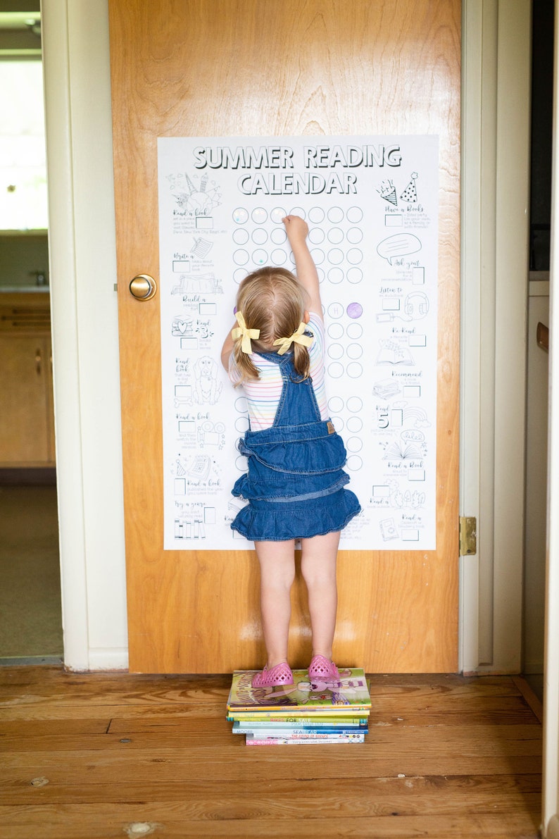 Op de afbeelding: Een jong meisje reikt naar een zomerleeskalenderposter op een deur. De poster heeft een raster met cirkels om in te kleuren terwijl het kind leest. Het meisje staat op een stapel boeken.