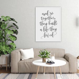 May include: A white framed print with black calligraphy that reads "and so together, they built a life they loved". The print is hanging on a white wall above a beige couch with a white throw pillow and a white coffee table with a vase of white flowers.