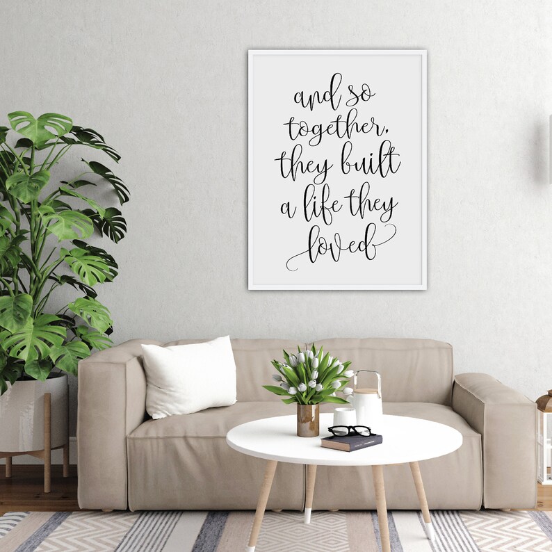 May include: A white framed print with black calligraphy that reads "and so together, they built a life they loved". The print is hanging on a white wall above a beige couch with a white throw pillow and a white coffee table with a vase of white flowers.
