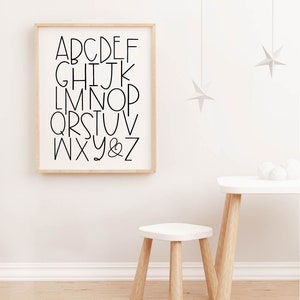 May include: A black and white alphabet poster with a simple, modern font. The poster is framed in a light wood frame and is hanging on a white wall. There is a small white table and stool in the foreground.
