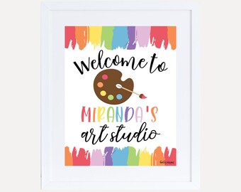 Può includere: Un cartello di benvenuto colorato per uno studio d'arte con un bordo di pennelli arcobaleno, una tavolozza di colori e il testo "Benvenuti allo studio d'arte di Miranda".