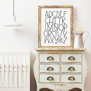 May include: A white dresser with six drawers and antique-style handles. A framed print with black lettering of the alphabet hangs above the dresser. A white lamp hangs from the ceiling above the dresser.