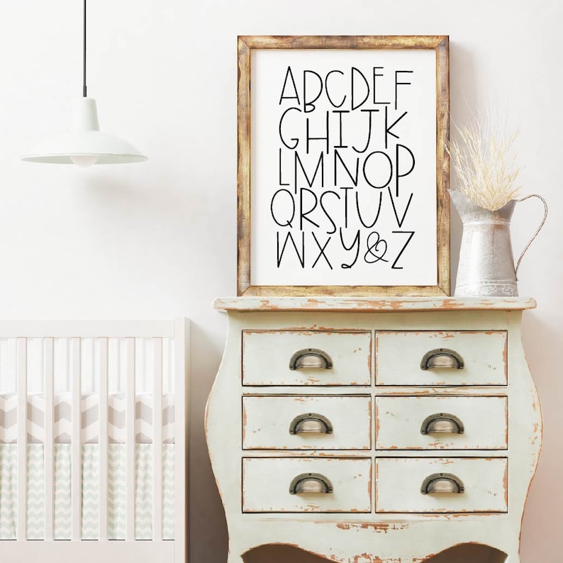 May include: A white dresser with six drawers and antique-style handles. A framed print with black lettering of the alphabet hangs above the dresser. A white lamp hangs from the ceiling above the dresser.