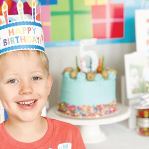Puede incluir: Un ni&ntilde;o peque&ntilde;o con una corona de papel azul y blanca con las palabras "&iexcl;Feliz cumplea&ntilde;os!" en ella. Est&aacute; sonriendo y mirando a la c&aacute;mara. Hay un pastel de cumplea&ntilde;os azul y blanco con chispas en el fondo.