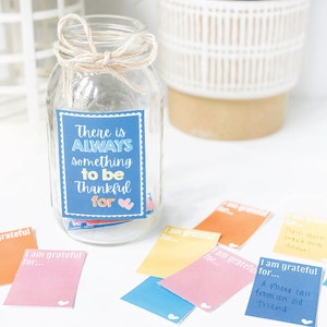 K&ouml;nnte beinhalten: Ein Glasgef&auml;&szlig; mit einem blauen und wei&szlig;en Schild, auf dem "There is ALWAYS something to be Thankful for." steht. Das Gef&auml;&szlig; ist mit kleinen bunten Papierschnipseln gef&uuml;llt, auf denen "I am grateful for..." steht.