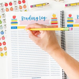 Pode incluir: Um di&aacute;rio de leitura com uma capa rosa e azul que apresenta uma pilha de livros com ma&ccedil;&atilde;s no topo. O di&aacute;rio tem uma linha amarela para a data e um espa&ccedil;o para notas. Uma m&atilde;o est&aacute; escrevendo no di&aacute;rio com um l&aacute;pis amarelo.