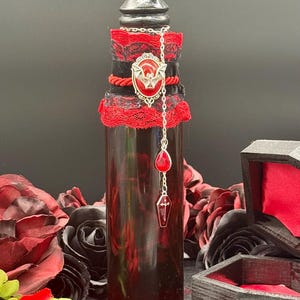 Peut inclure: Une bouteille décorative remplie d'un liquide rouge foncé, avec un bouchon noir et un col en dentelle rouge et noire. Une chaîne argentée avec des breloques rouges, dont un cercueil, pend du col. La bouteille est entourée de roses rouges et d'une petite boîte en forme de cercueil.