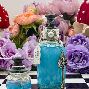 May include: Two decorative glass bottles with blue liquid, adorned with lace, pearls, and key charms. One bottle has a tag that reads "DRINK ME!". The bottles are set against a backdrop of colorful flowers, evoking a whimsical theme.