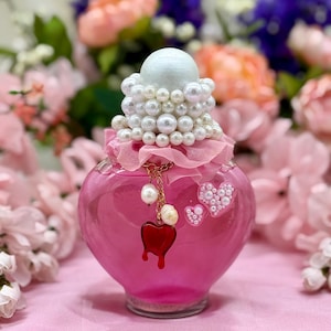 May include: A pink, heart-shaped glass bottle with a white stopper adorned with pearls. A red heart charm hangs from the neck, and pearl-studded hearts decorate the bottle. The bottle is filled with a pink liquid, and is surrounded by flowers.