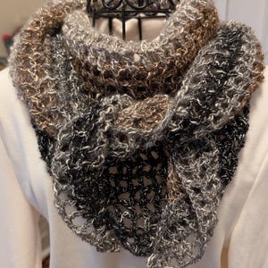 May include: A handmade, open-weave cowl scarf in shades of brown, gray, and black. The scarf is crocheted with a textured yarn, creating a soft and warm accessory. The cowl is displayed on a mannequin, showcasing its shape and drape.