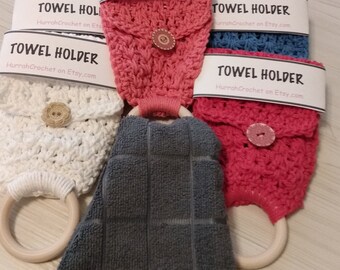May include: A set of crocheted towel holders in various colors, including white, pink, and blue. Each holder features a button and a loop for hanging on a towel rack. The holders are displayed on a wooden surface with a gray towel.