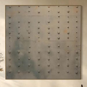 May include: A large, square, grey metal wall art piece with a grid of circular rivets. Below, a natural wood bench with a vase of dried flowers and a black decorative bowl. The room has a neutral colour palette.