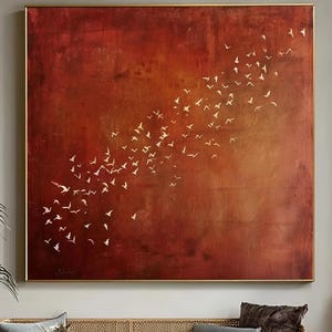 Könnte beinhalten: Ein großes, quadratisches abstraktes Gemälde mit tiefrotem und goldenem Farbverlauf im Hintergrund. Weiße Vogelsilhouetten fliegen über die Leinwand. Das Gemälde hängt über einem grauen Sofa mit Holzrahmen, einem goldenen Couchtisch und einem gemusterten Sessel.