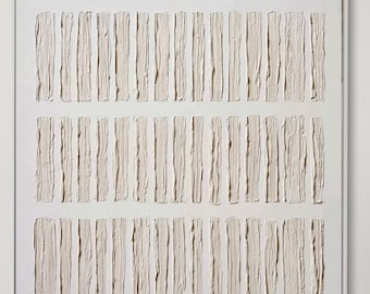 Op de afbeelding: Groot, ingelijst abstract kunstwerk met een textuur, neutraal getint ontwerp. Het kunstwerk toont verticale rechthoekige vormen gerangschikt in drie rijen. Het hangt boven een witte bank met kussens, een rond tapijt en een houten bijzettafel.