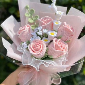 Puede incluir: Un ramo de rosas rosas de ganchillo, margaritas blancas y vegetación envuelto en papel de seda rosa y una cinta transparente. Las flores hechas a mano están en plena floración, creando un arreglo floral delicado y encantador.