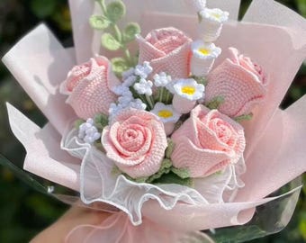 May include: A bouquet of crocheted pink roses, white daisies, and greenery wrapped in pink tissue paper and a sheer ribbon. The handmade flowers are in full bloom, creating a delicate and charming floral arrangement.