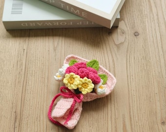 May include: A handmade crochet bouquet featuring pink, yellow, and white flowers with green leaves. The bouquet is wrapped in pink yarn and tied with a bow. The bouquet is placed on a wooden surface.