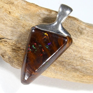 May include: A triangular pendant with a silver bail. The pendant features a polished, dark brown stone with iridescent flashes of colour. The stone is set in a silver bezel. The pendant is displayed on a piece of driftwood.