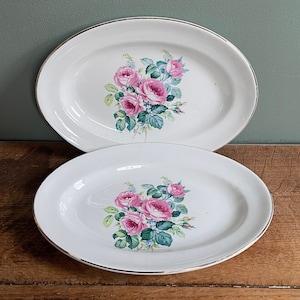 May include: Two white oval serving dishes with a gold rim. Both dishes feature a floral design with pink roses, green leaves, and blue accents.