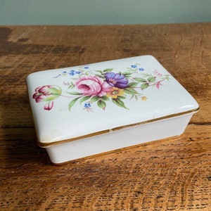 May include: A white rectangular ceramic trinket box with a gold trim and a floral design on the lid. The flowers are pink, purple, yellow, and blue.