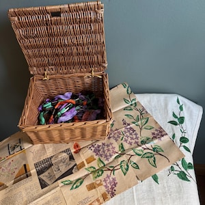 May include: A woven wicker basket, lid open, containing colourful embroidery threads. Next to the basket is a vintage linen with embroidered lilac and leaf designs. A vintage pattern is also visible.